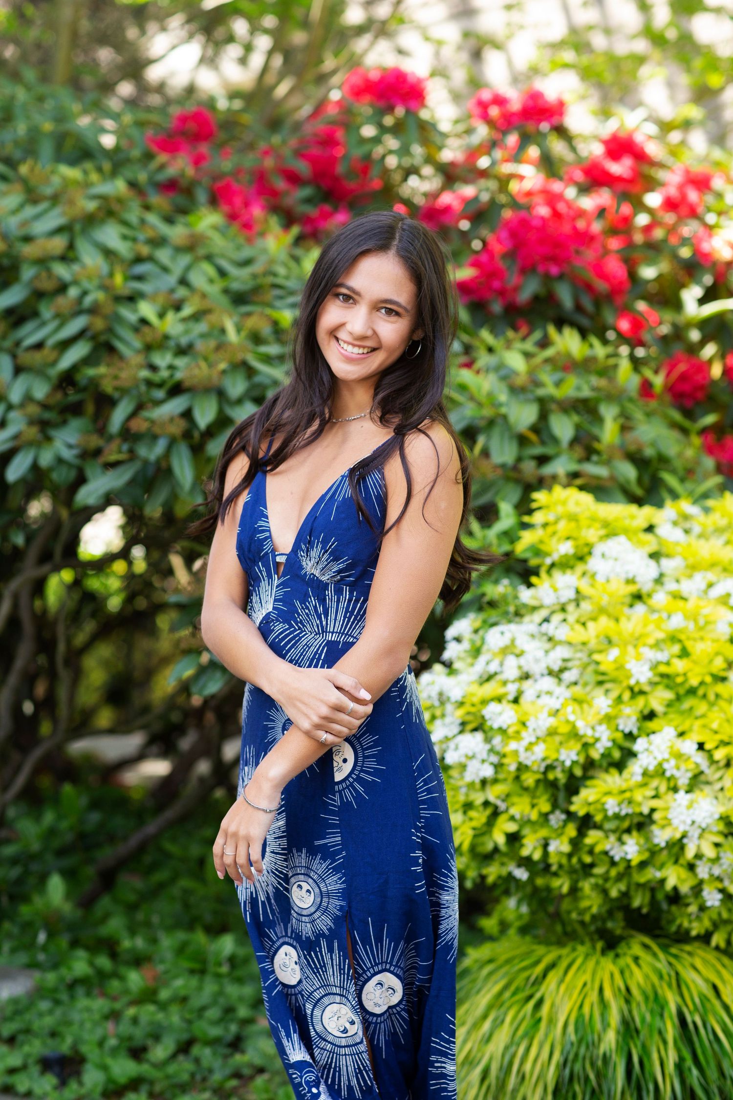 Young woman in blue floral maxi dress smiling in garden with red and white blooming flowers.
