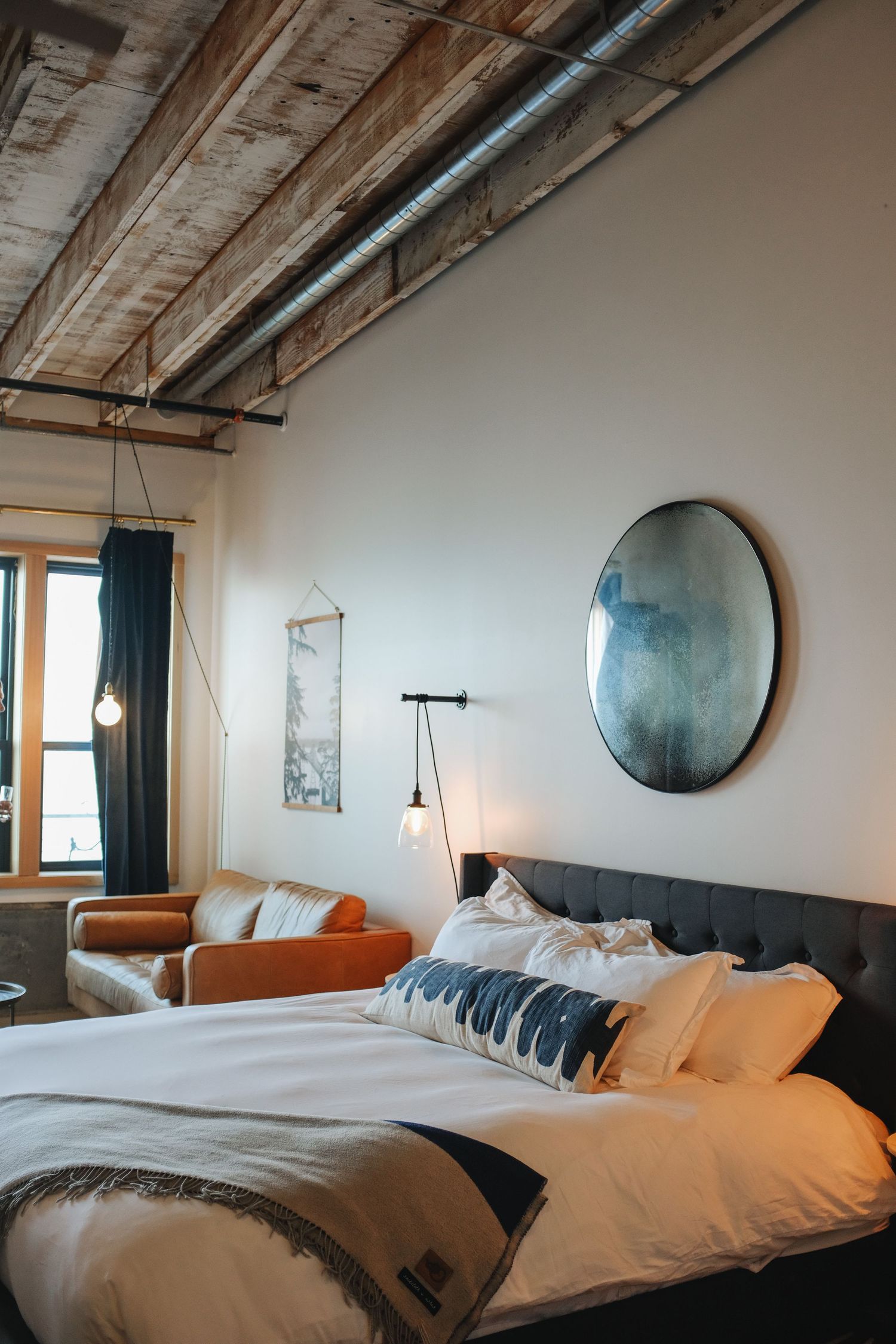 A stylish hotel bedroom with rustic wood ceiling, industrial lighting, round mirror, and neutral bedding.