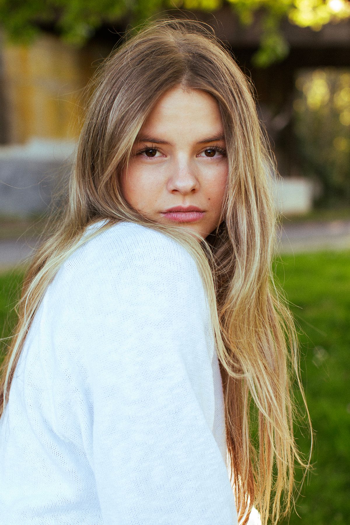 Young girl with long blonde hair wearing a white sweater, looking over her shoulder outdoors.