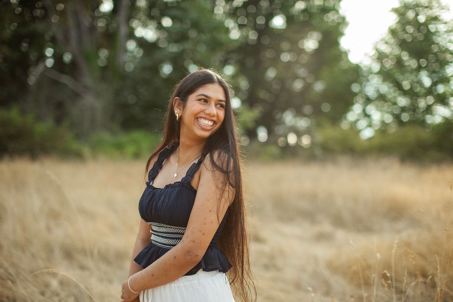 Smiling young woman with long dark hair in black top and white skirt standing in golden field.