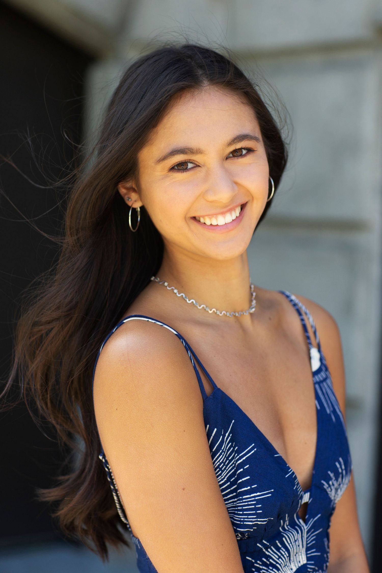 Smiling young woman with long dark hair wearing a blue floral dress and gold hoop earrings outdoors.