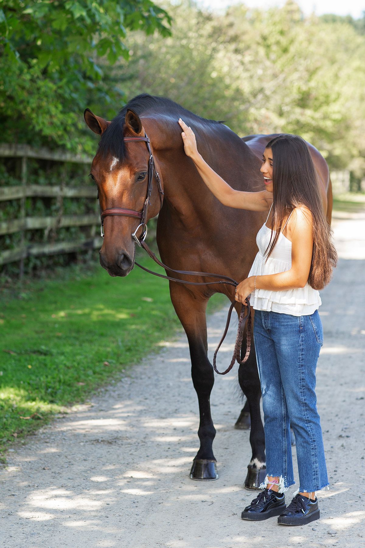 Young woman in jeans petting a bay horse on a sunny gravel path near a wooden fence.