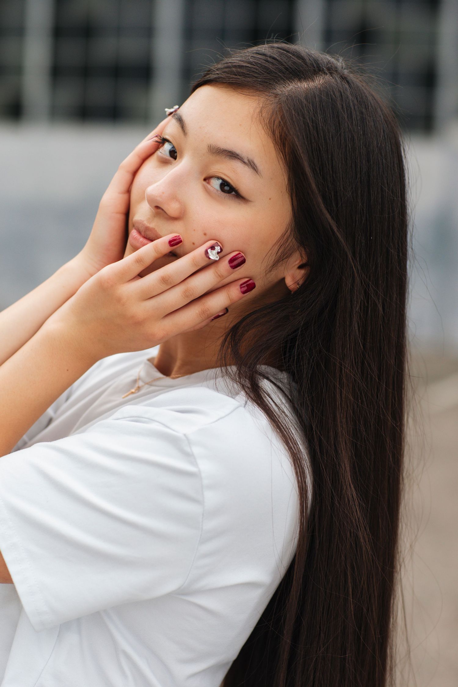 Young woman with dark red nails gently touching her face, wearing white top in outdoor setting.