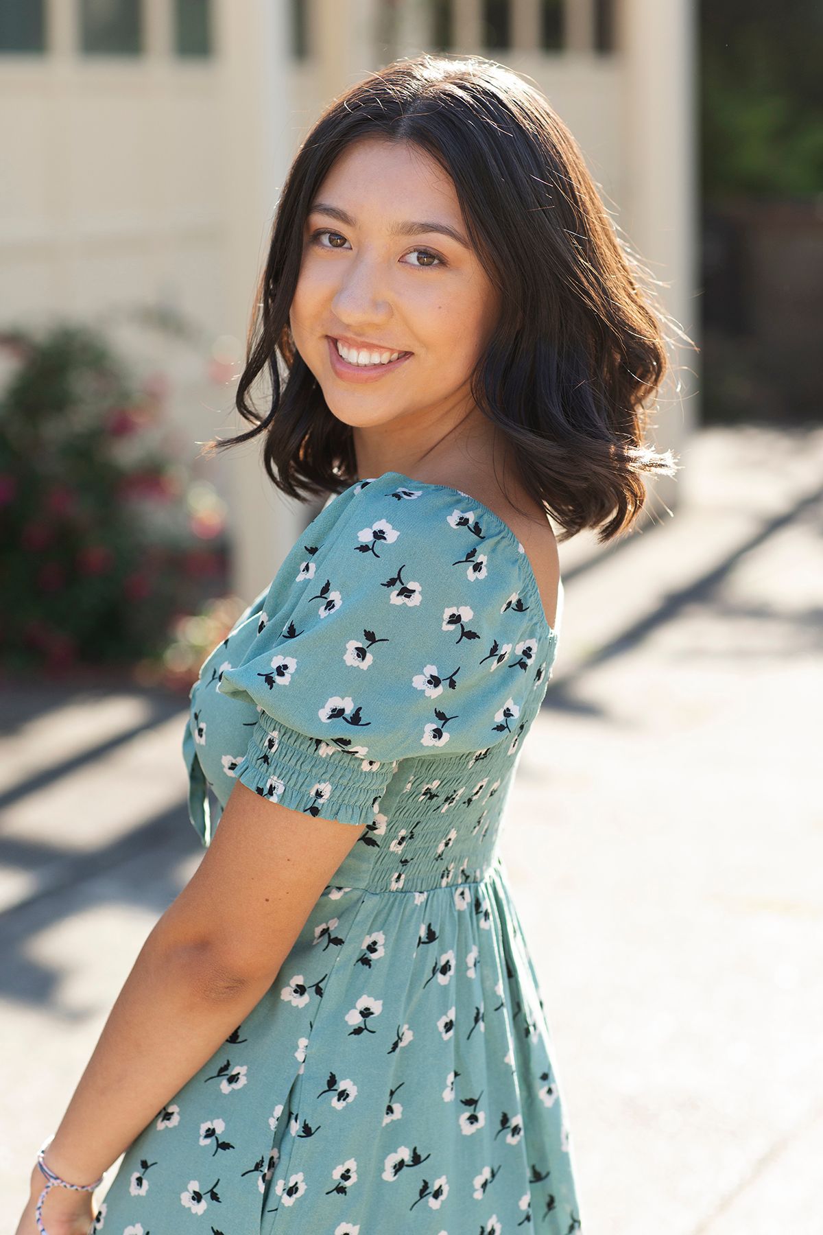 Young woman smiling in a green floral off-shoulder dress, posing outdoors in warm sunlight.
