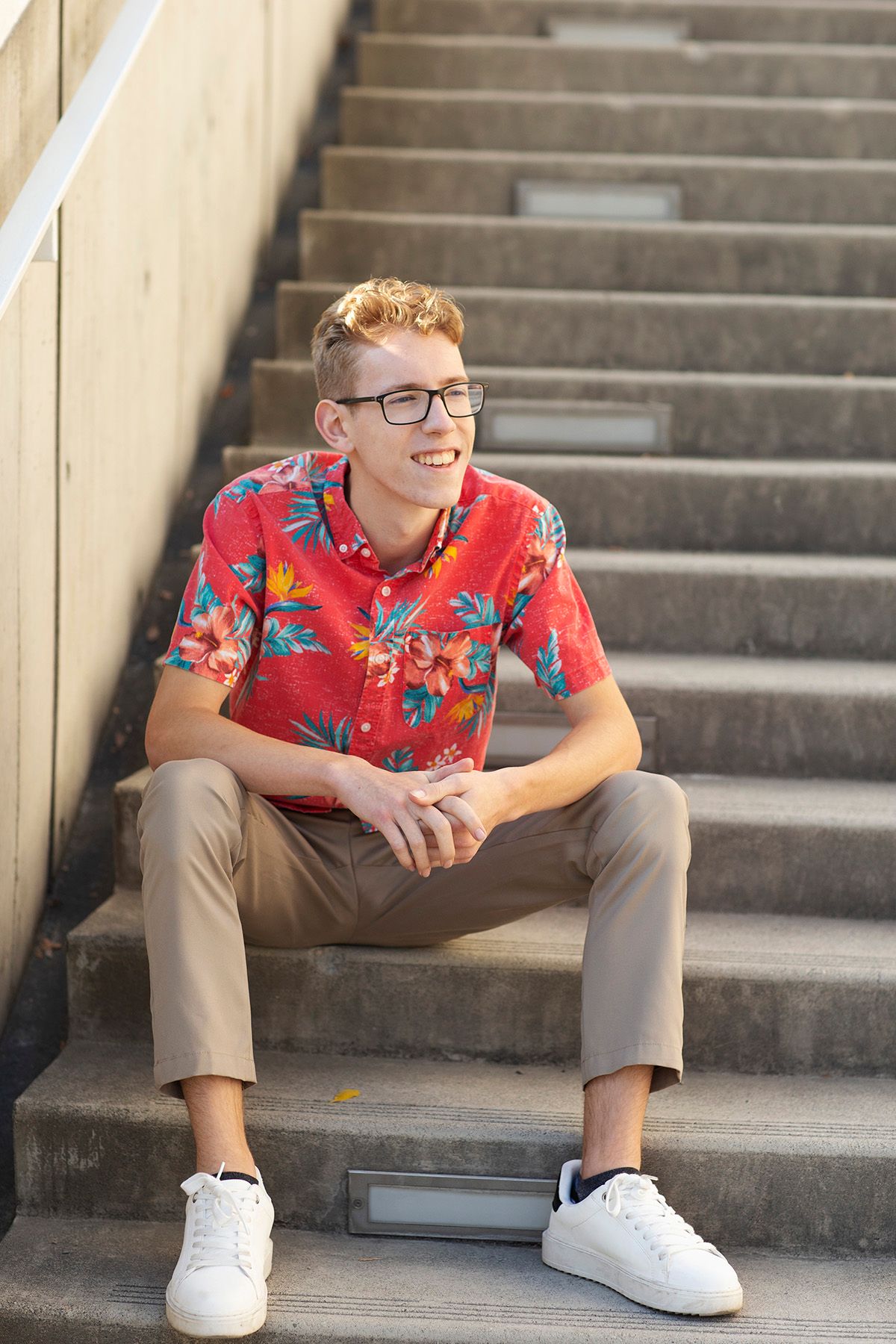 Young man in red floral shirt and glasses sitting on concrete steps, smiling outdoors.