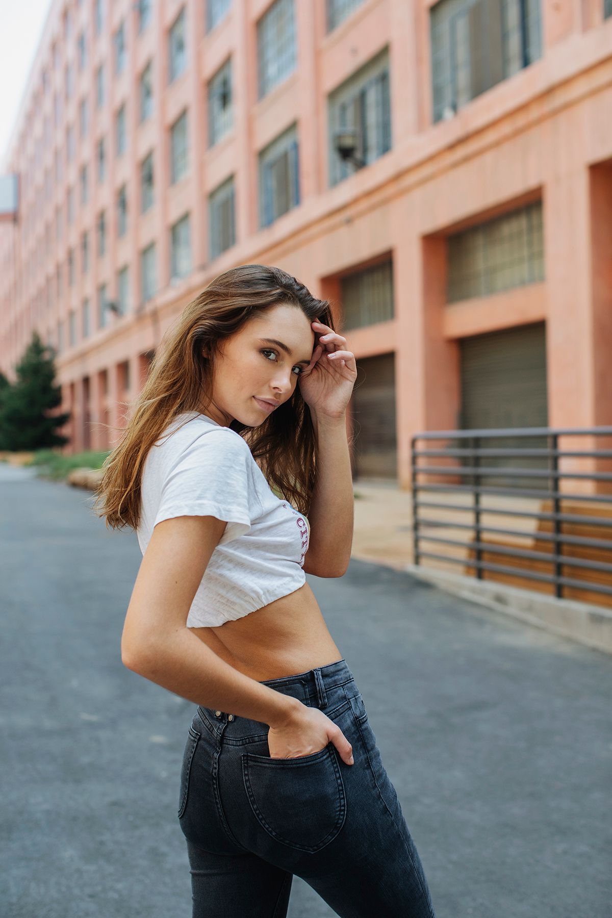 Young woman in white crop top and dark jeans posing outdoors near urban brick building.