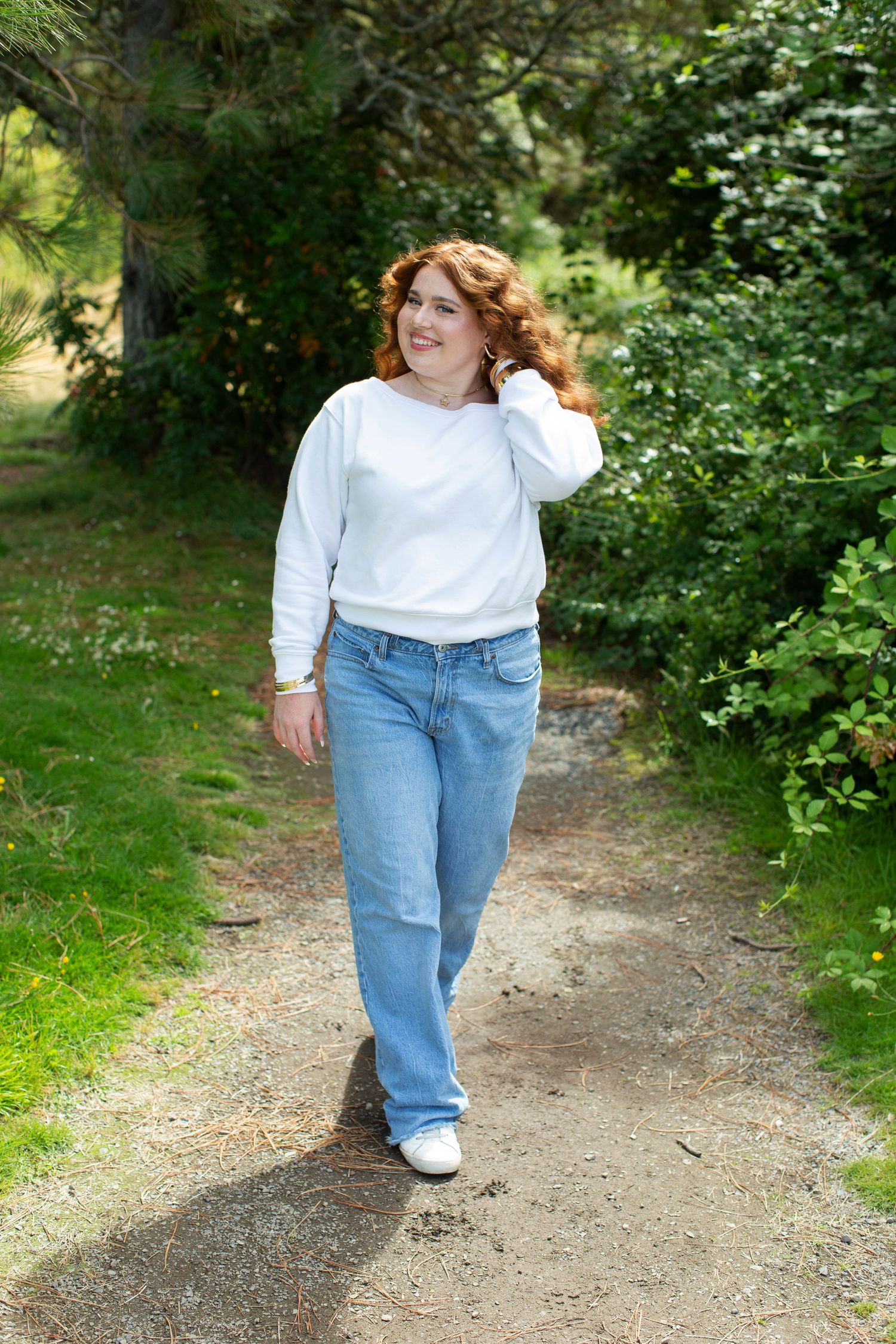 Young woman with red curly hair in white sweatshirt and jeans walking on a scenic garden path.