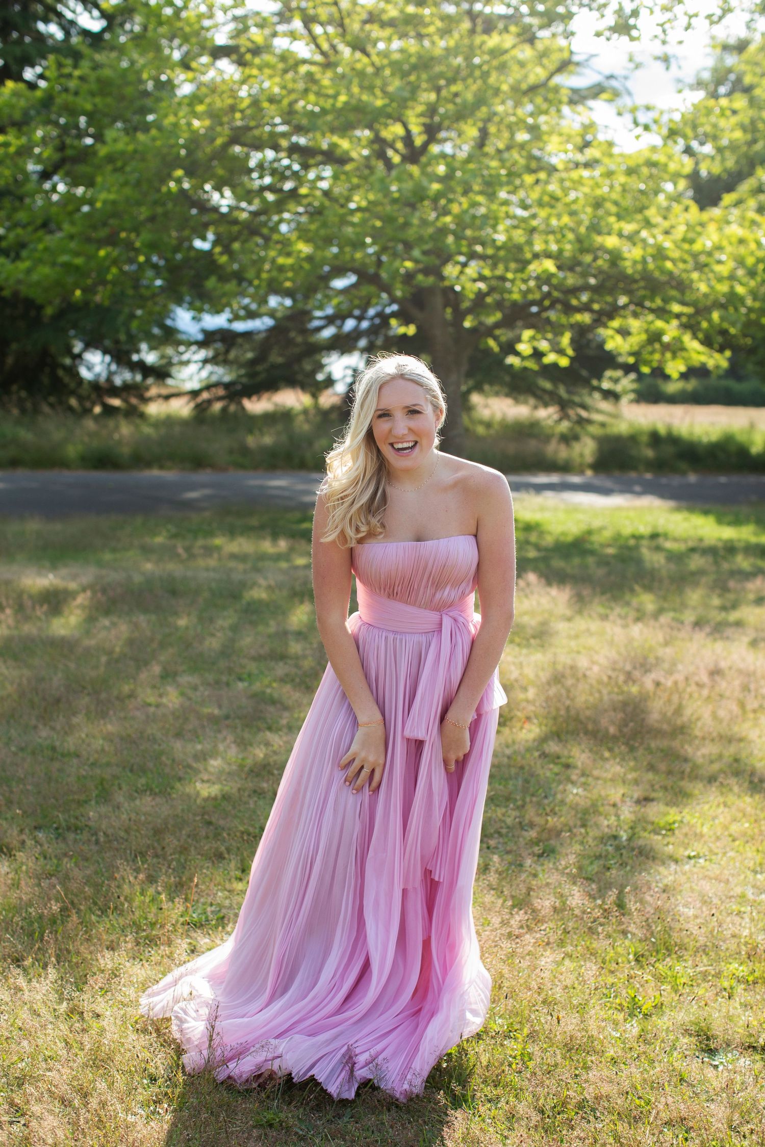 Blonde woman laughing in a flowing strapless pink tulle gown standing on a grassy field with trees behind.