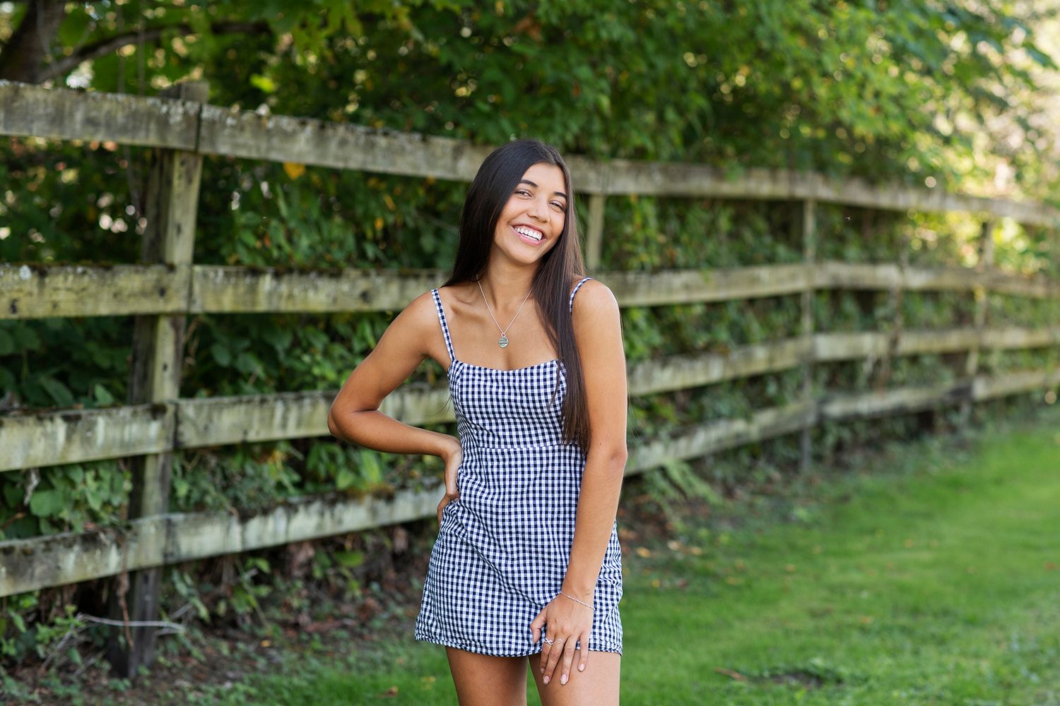 Smiling young woman in gingham dress posing outdoors by a rustic wooden fence on a sunny day.