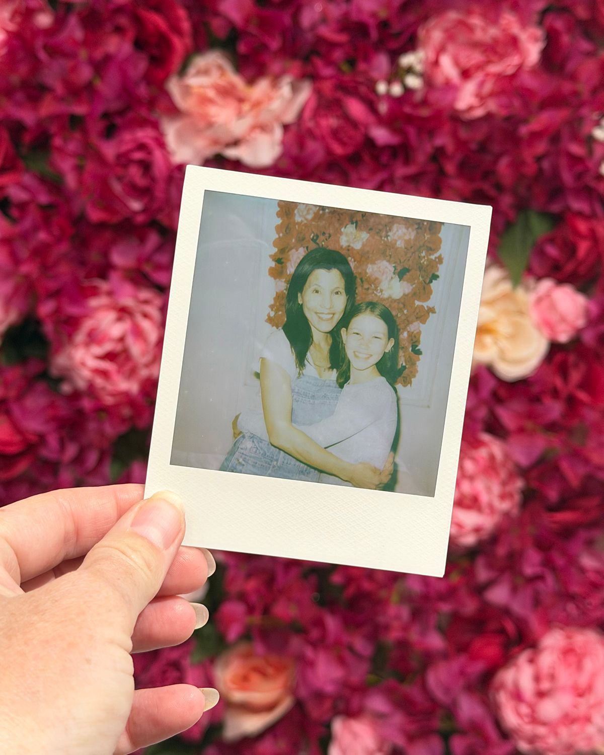 Hand holding a Polaroid photo of two women smiling, set against a vibrant pink floral wall backdrop.