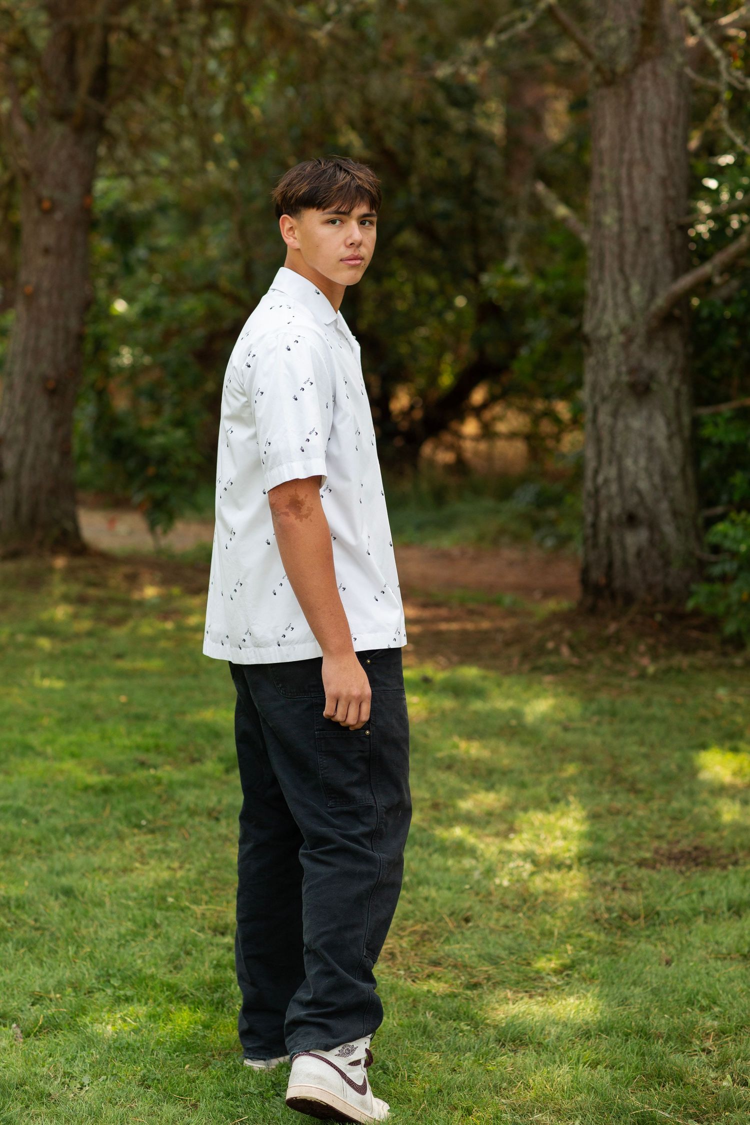 Teen boy in white patterned shirt and black pants standing on green grass among trees.