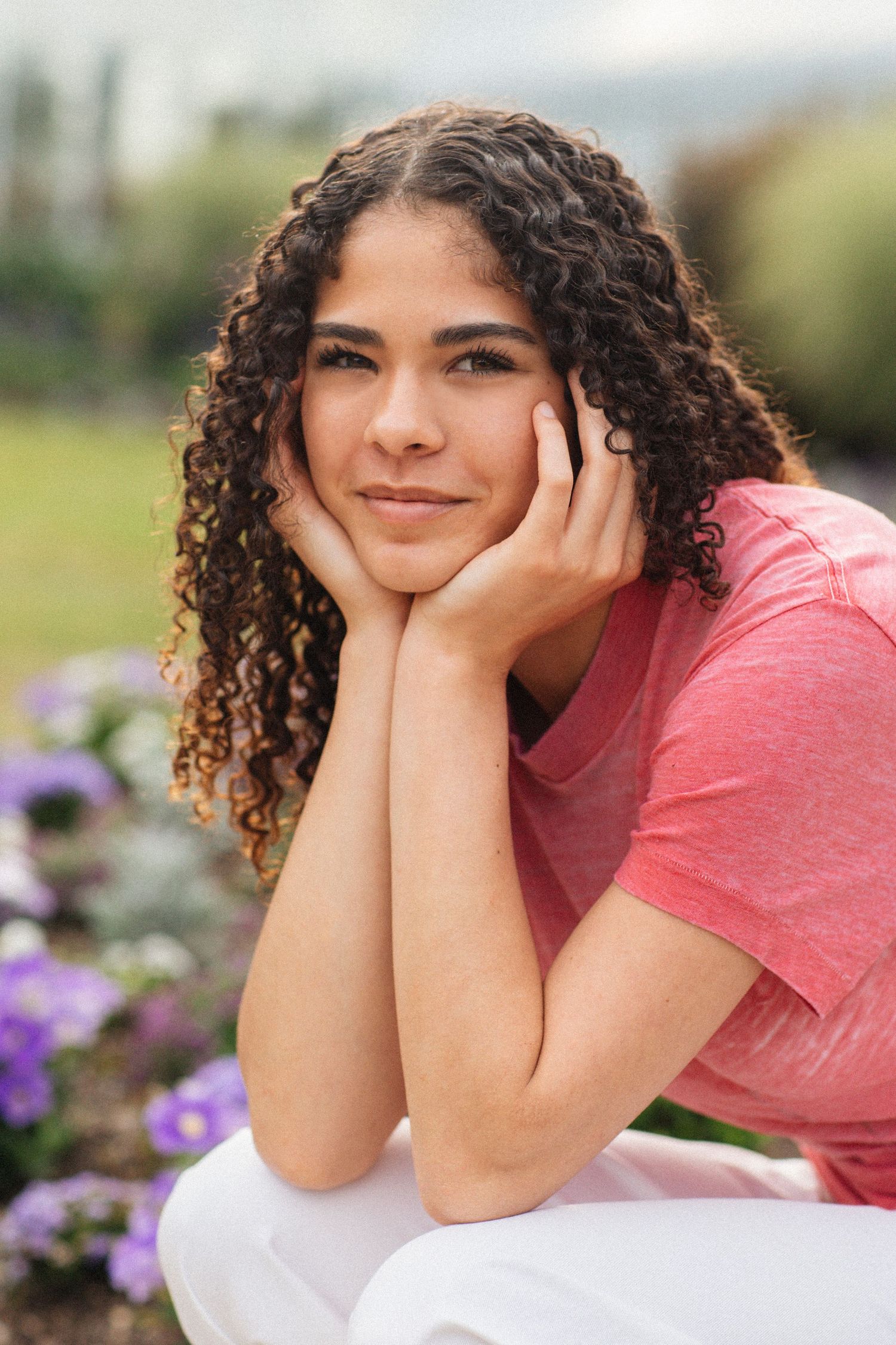 Smiling young woman with curly hair resting chin on hands in outdoor garden with purple flowers.