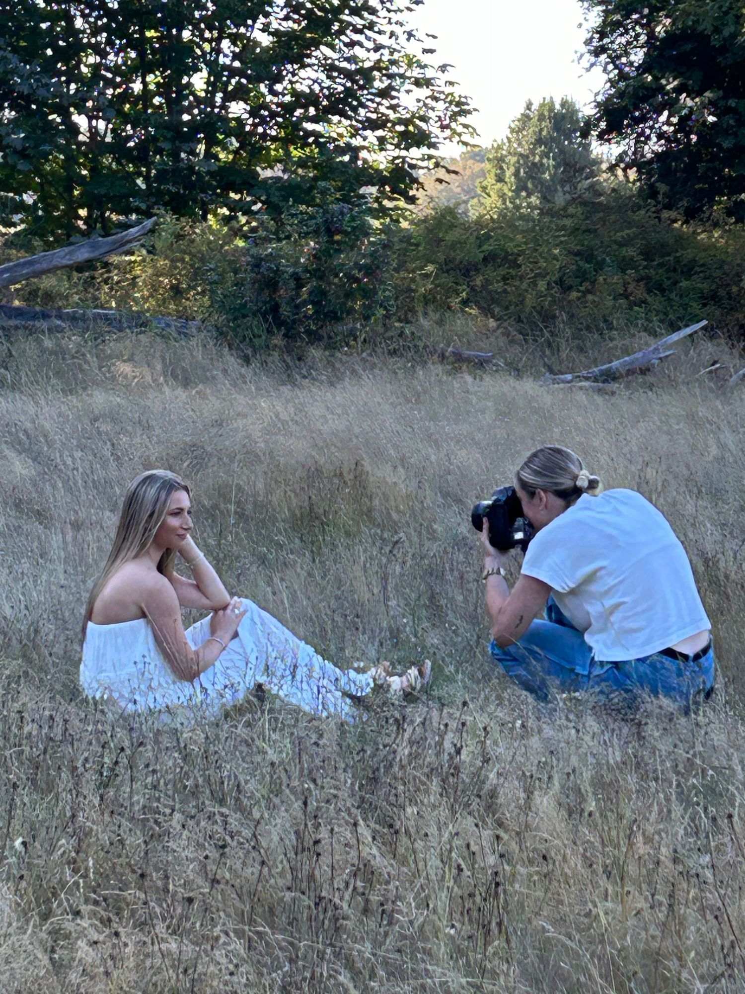 Photographer captures woman in white dress sitting in tall golden grass field surrounded by trees.
