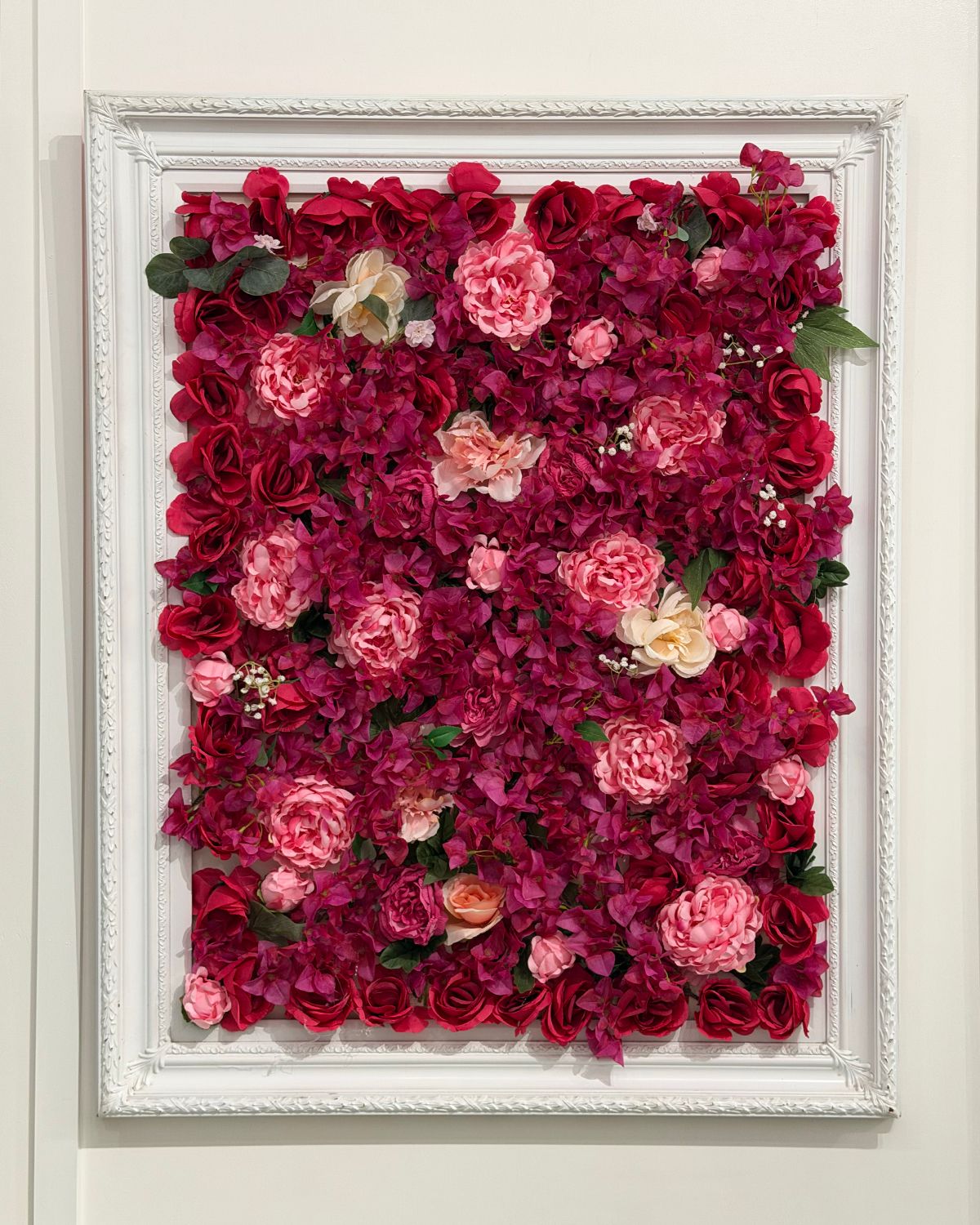 A decorative white framed wall art piece filled with vibrant red, pink, and cream artificial flowers.
