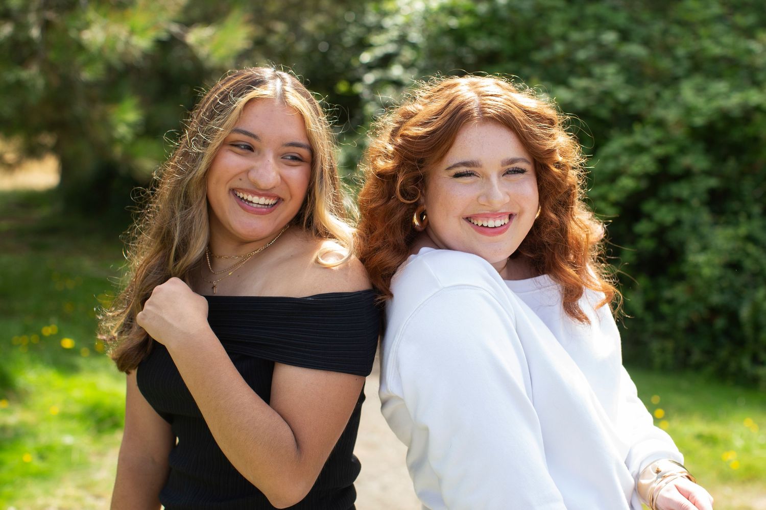 Two smiling young women posing together outdoors, one in black off-shoulder top and one in white jacket.