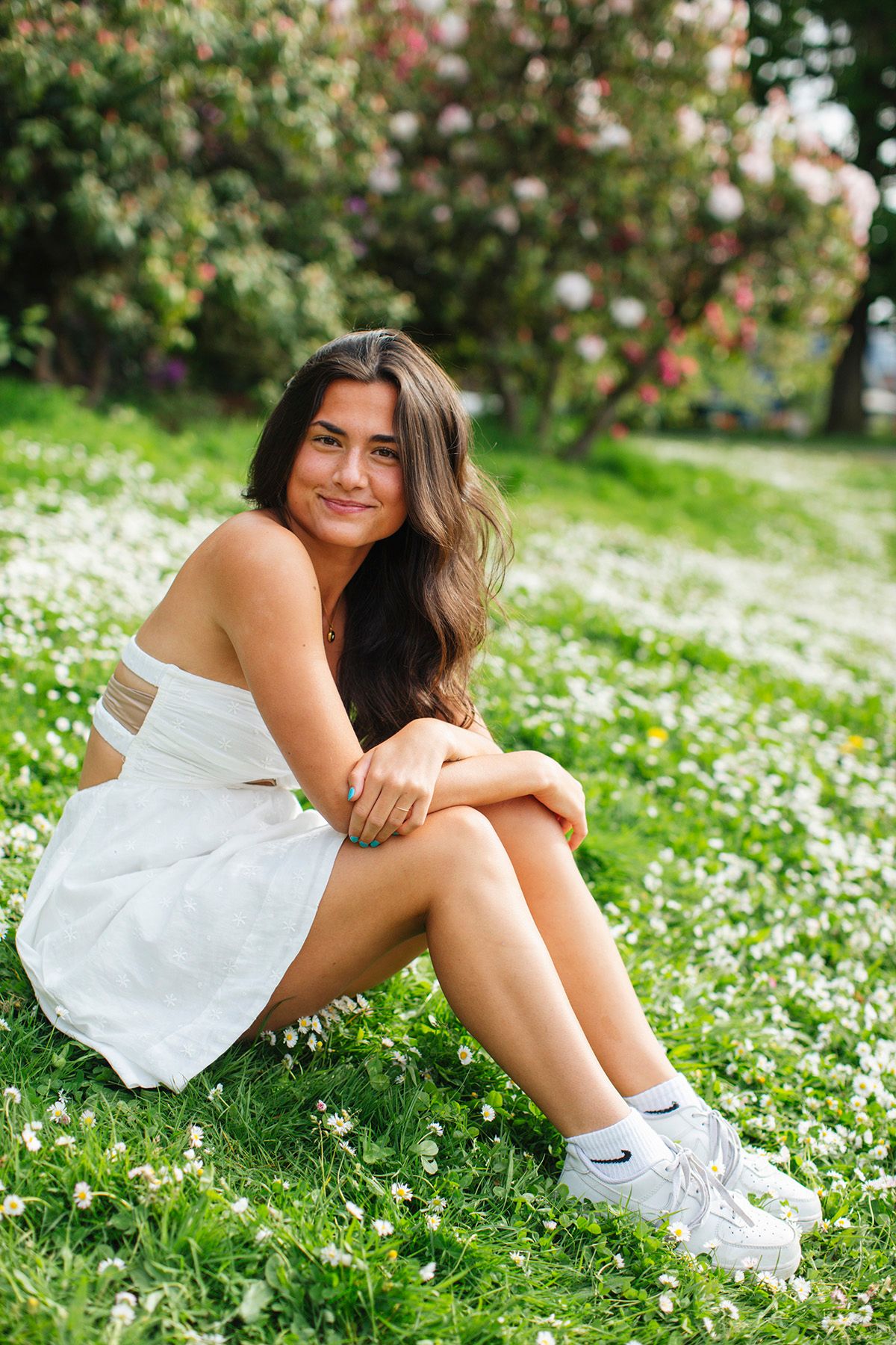 Young woman in white dress sitting on grass field with wildflowers, smiling outdoors near blooming trees.