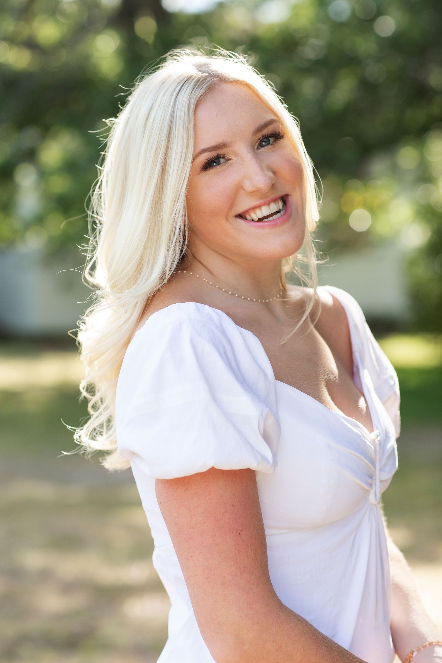 Smiling blonde woman in a white puff-sleeve dress posing outdoors in soft natural sunlight.