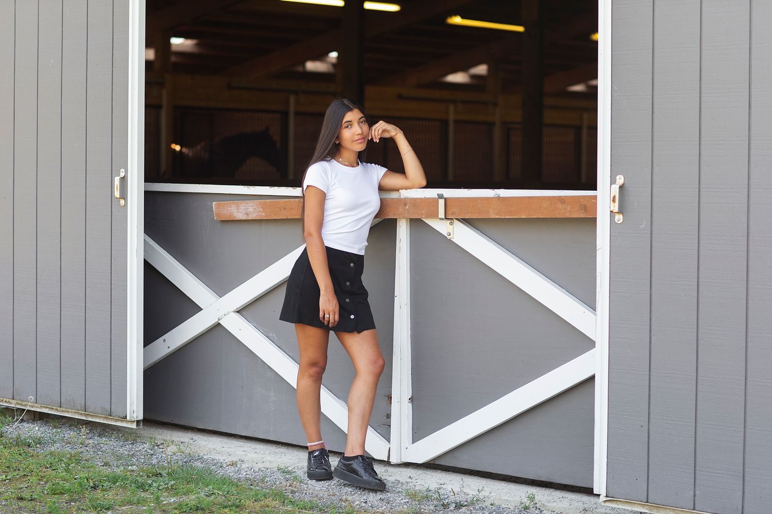 Young woman in white tee and black skirt posing confidently against a rustic barn door outdoors.