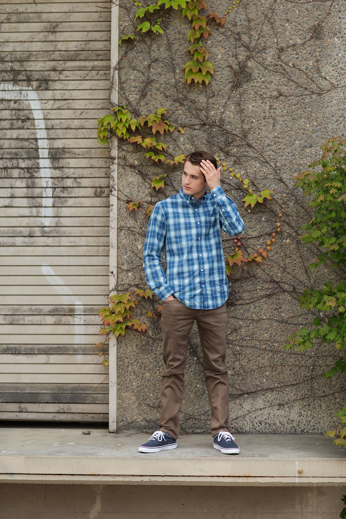 Young man in blue plaid shirt and khaki pants leaning against a stone wall with ivy, looking away thoughtfully.