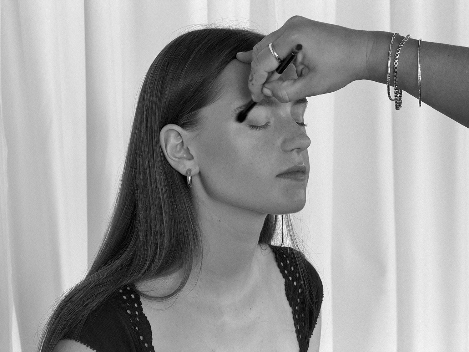 Black and white photo of a woman having eye makeup applied by a makeup artist with a brush.
