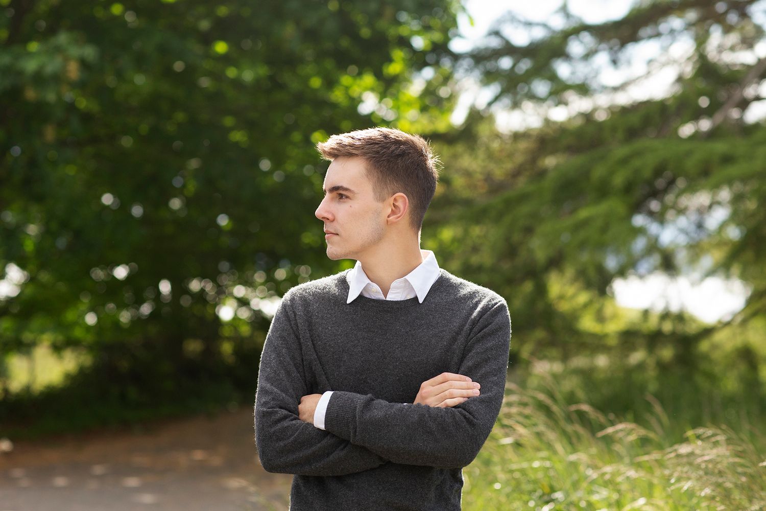 Young man in gray sweater stands outdoors with arms crossed, looking away amid lush green trees.