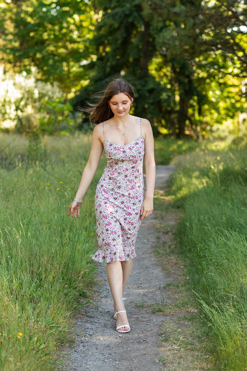 Young woman in floral sundress walking on a path surrounded by lush green trees and grass on a sunny day.