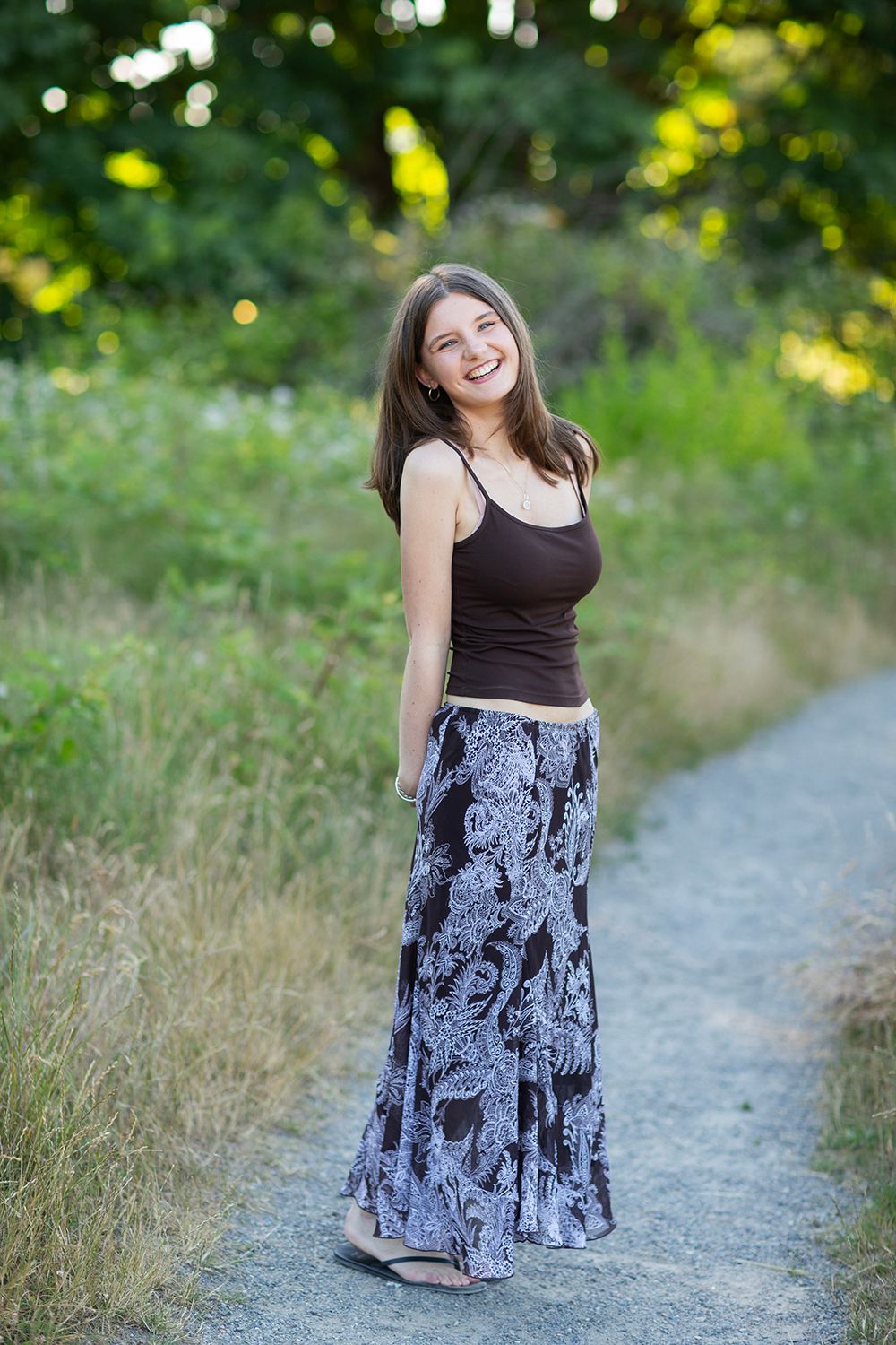 Smiling young woman in brown tank top and floral maxi skirt standing on a path surrounded by lush greenery.