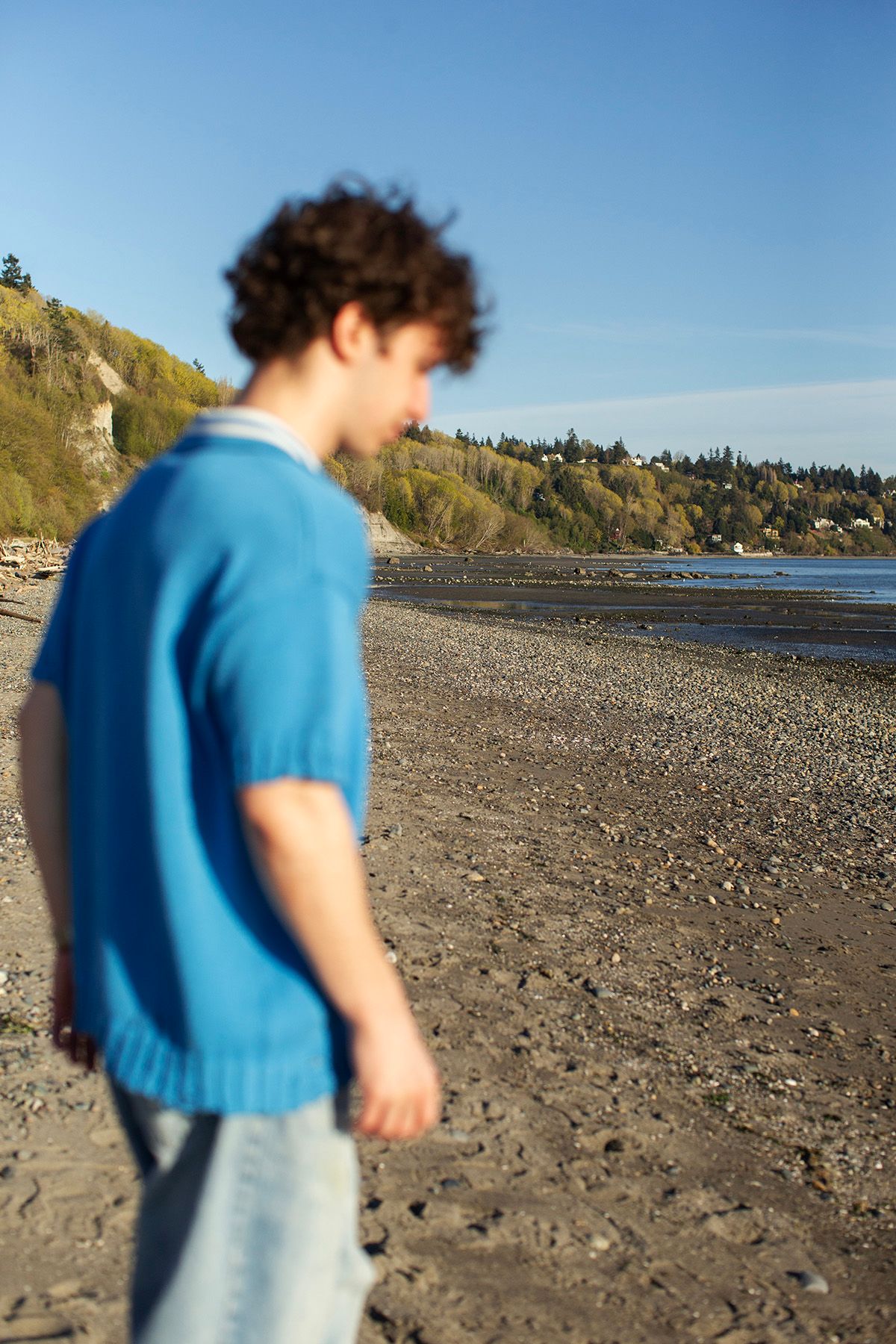 Young man in blue sweater walking on a rocky beach with trees and blue sky in background.