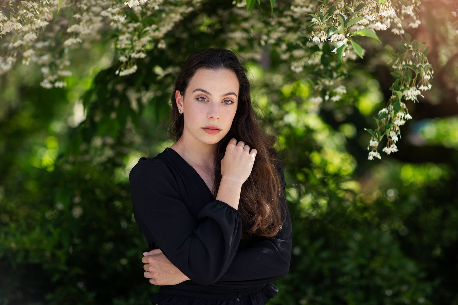 Young woman in black dress poses gracefully among lush green foliage and white blooming flowers outdoors.