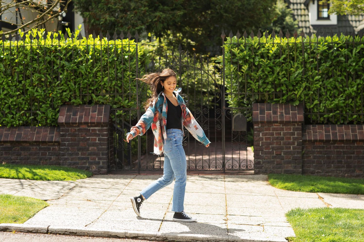 Young woman in colorful patterned jacket walking playfully along a sunny suburban sidewalk.