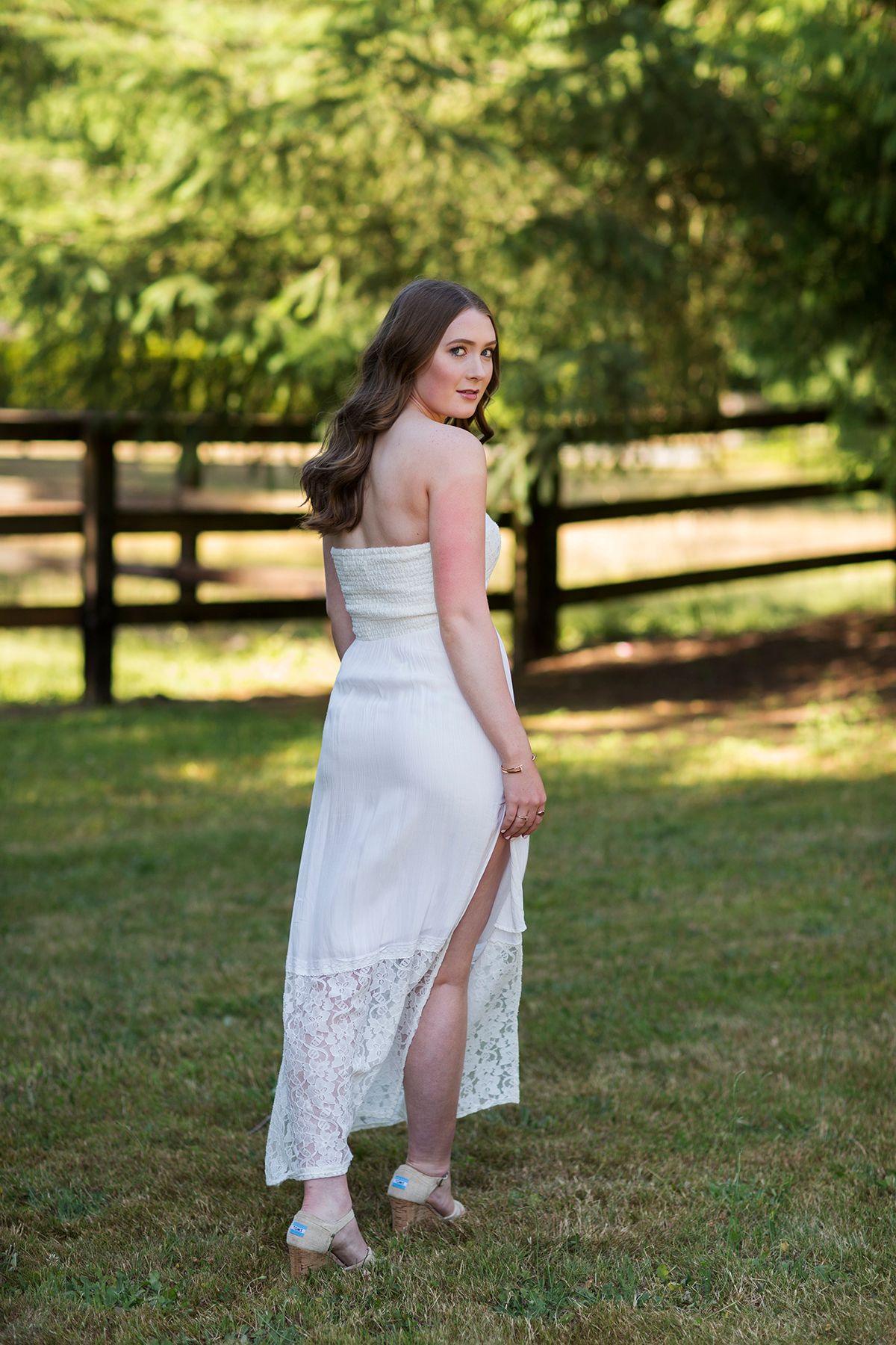 Young woman in white strapless lace dress poses outdoors on green lawn with wooden fence and trees behind her.