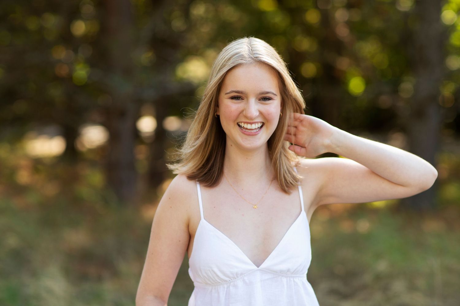 Young blonde woman in white sundress smiling outdoors with trees in background on sunny day.