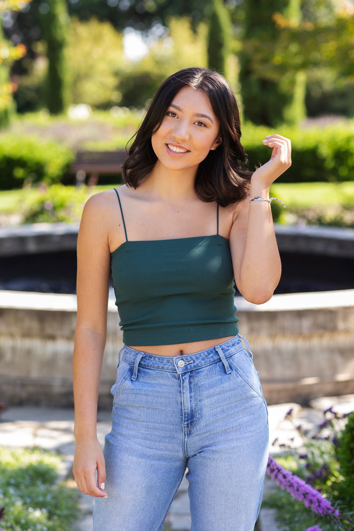 Young woman in green crop top and jeans smiling outdoors near a garden fountain.