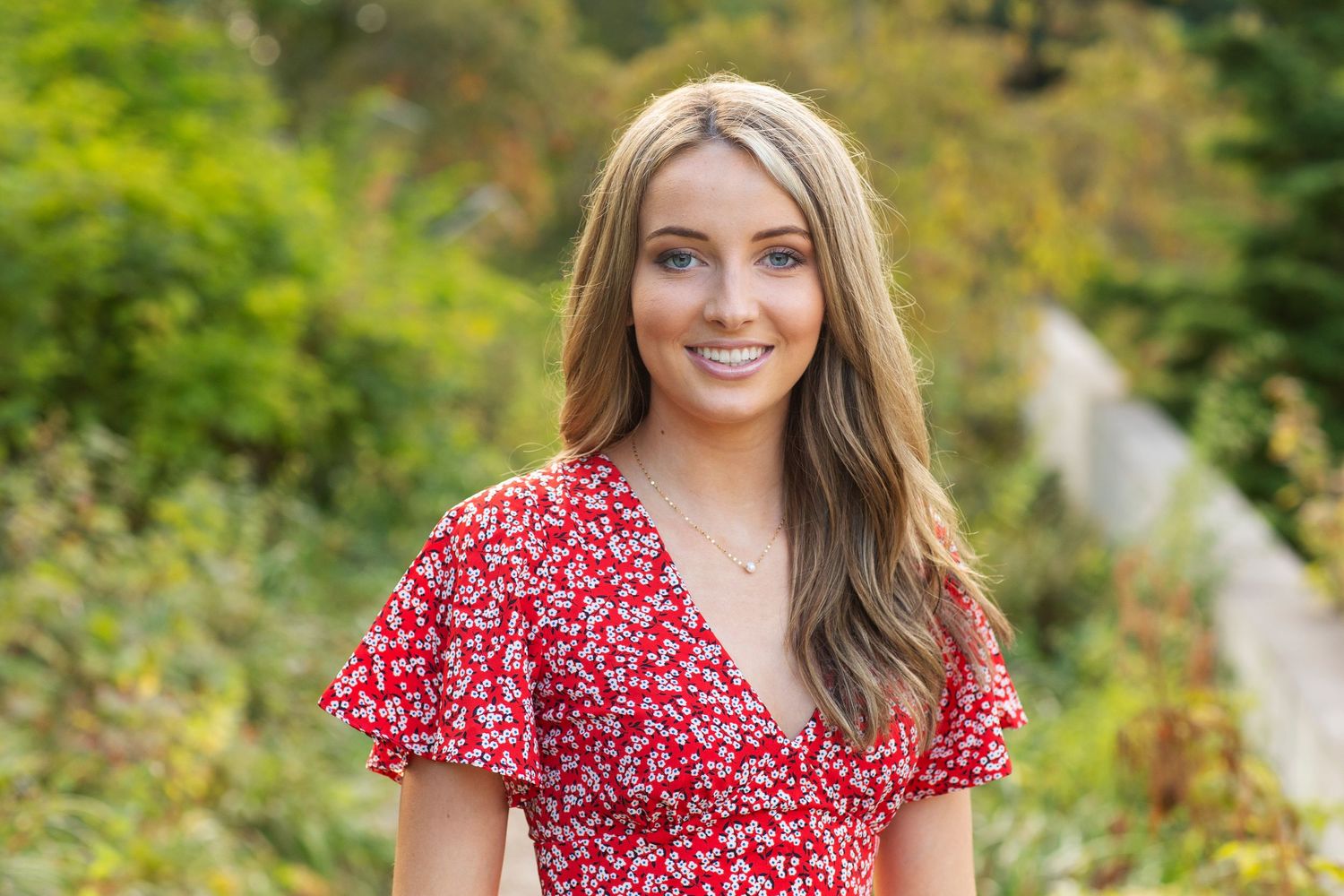 Smiling young woman in red floral dress posing outdoors among green foliage.