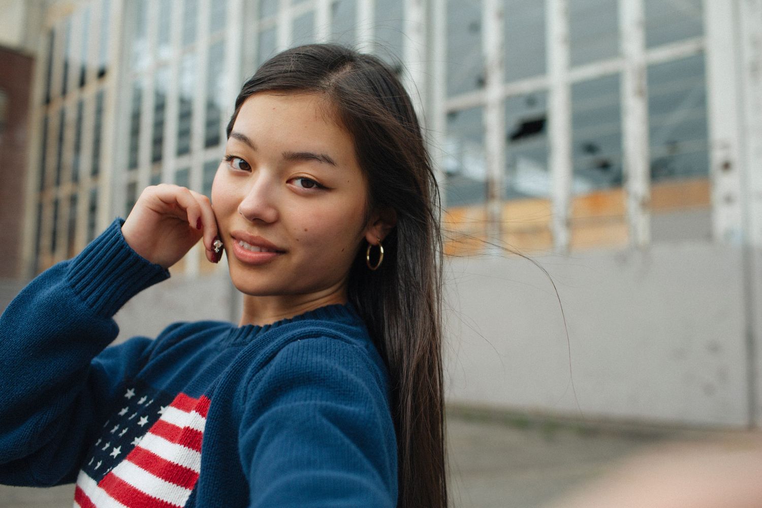 Young woman smiling softly in a navy American flag sweater, posing outdoors near an urban building.