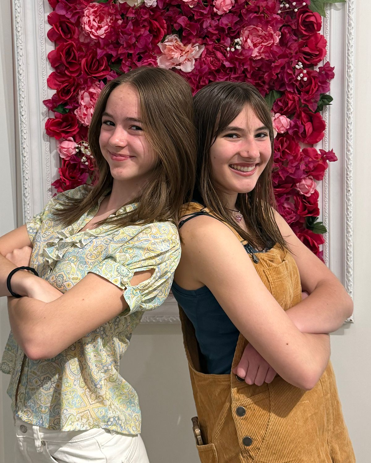 Two teenage girls posing back-to-back with arms crossed in front of a red floral wall backdrop.