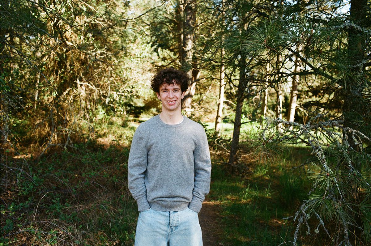 Young man in gray sweater and light jeans smiling on a forest path surrounded by green trees.