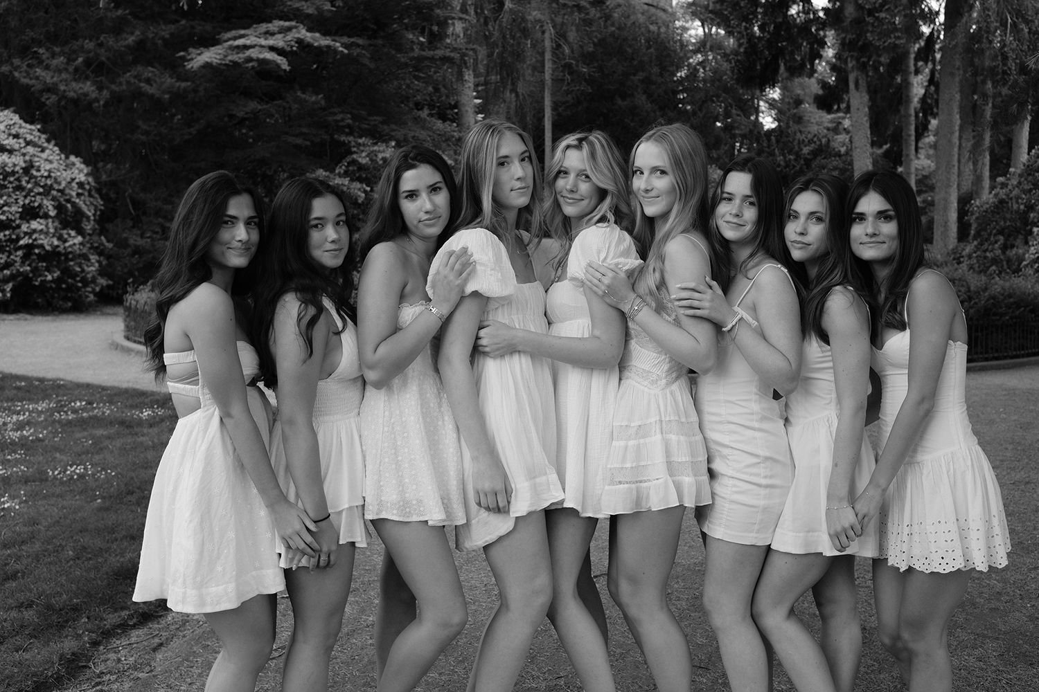 Group of nine young women posing together outdoors in white dresses for a black and white photo.
