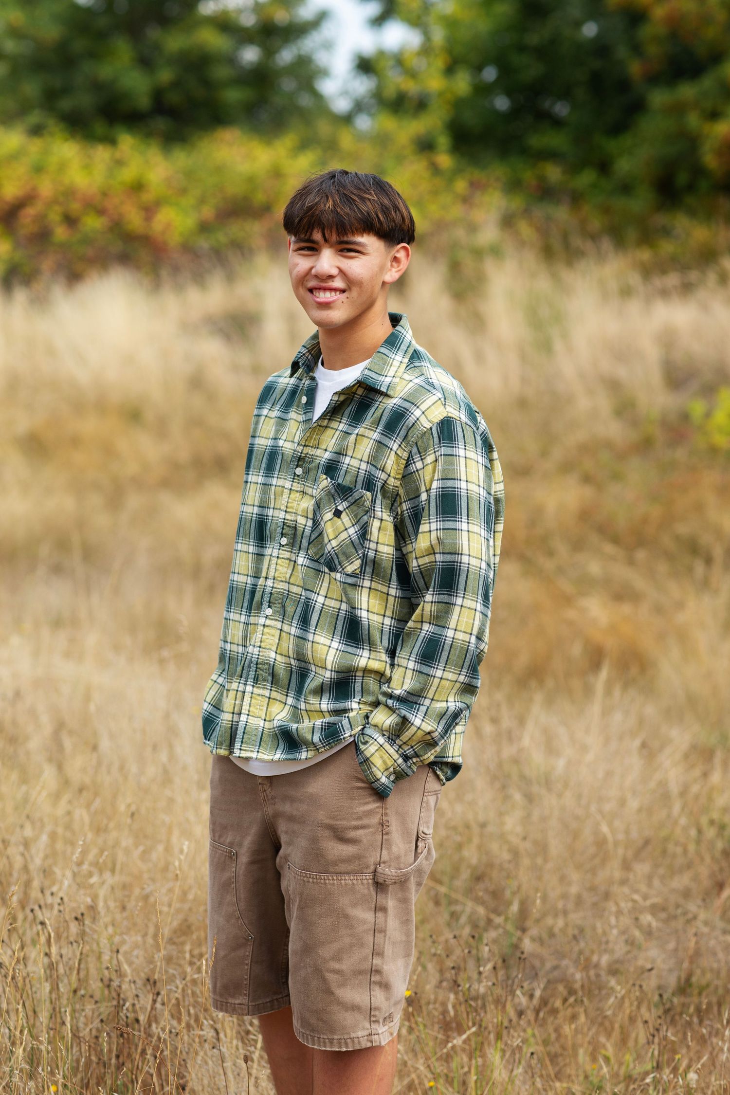 Young man in green plaid flannel shirt smiling in golden field with autumn foliage.