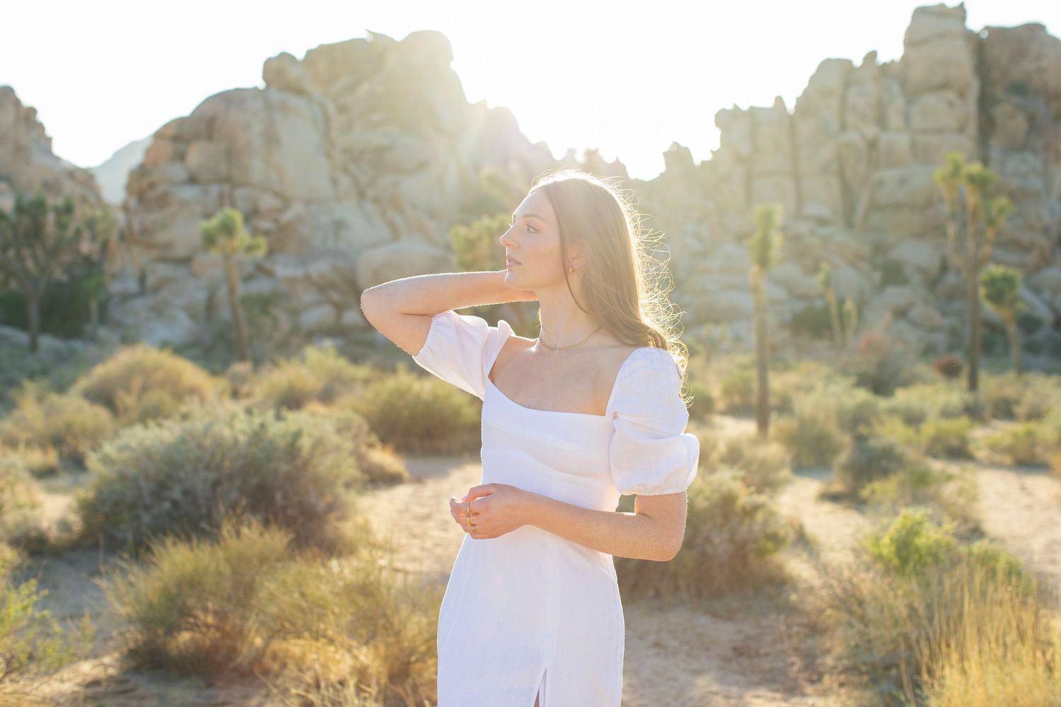 Young woman in white dress posing in sunlit desert landscape with rocky formations and Joshua trees.