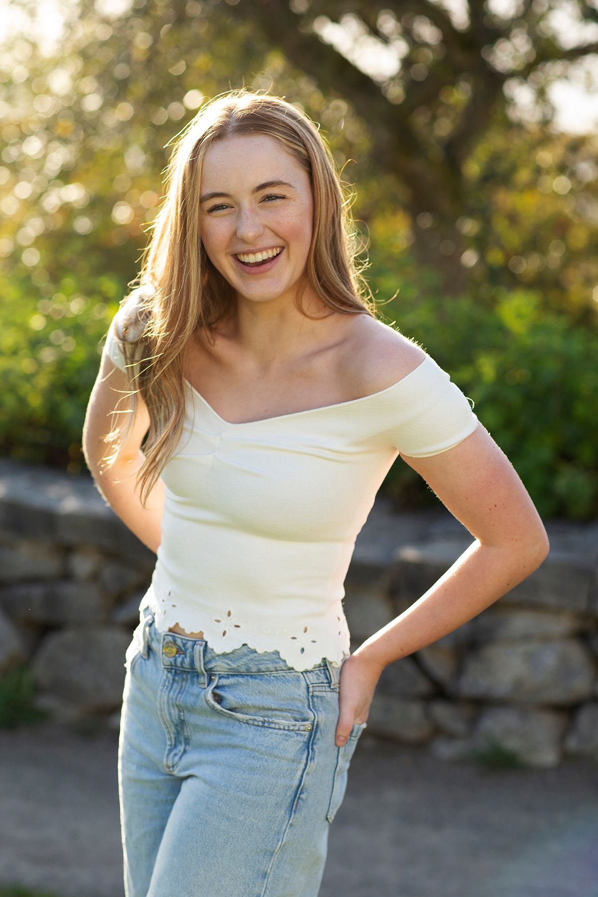 Smiling young woman with blonde hair wearing white off-shoulder top and jeans posing outdoors in golden sunlight.