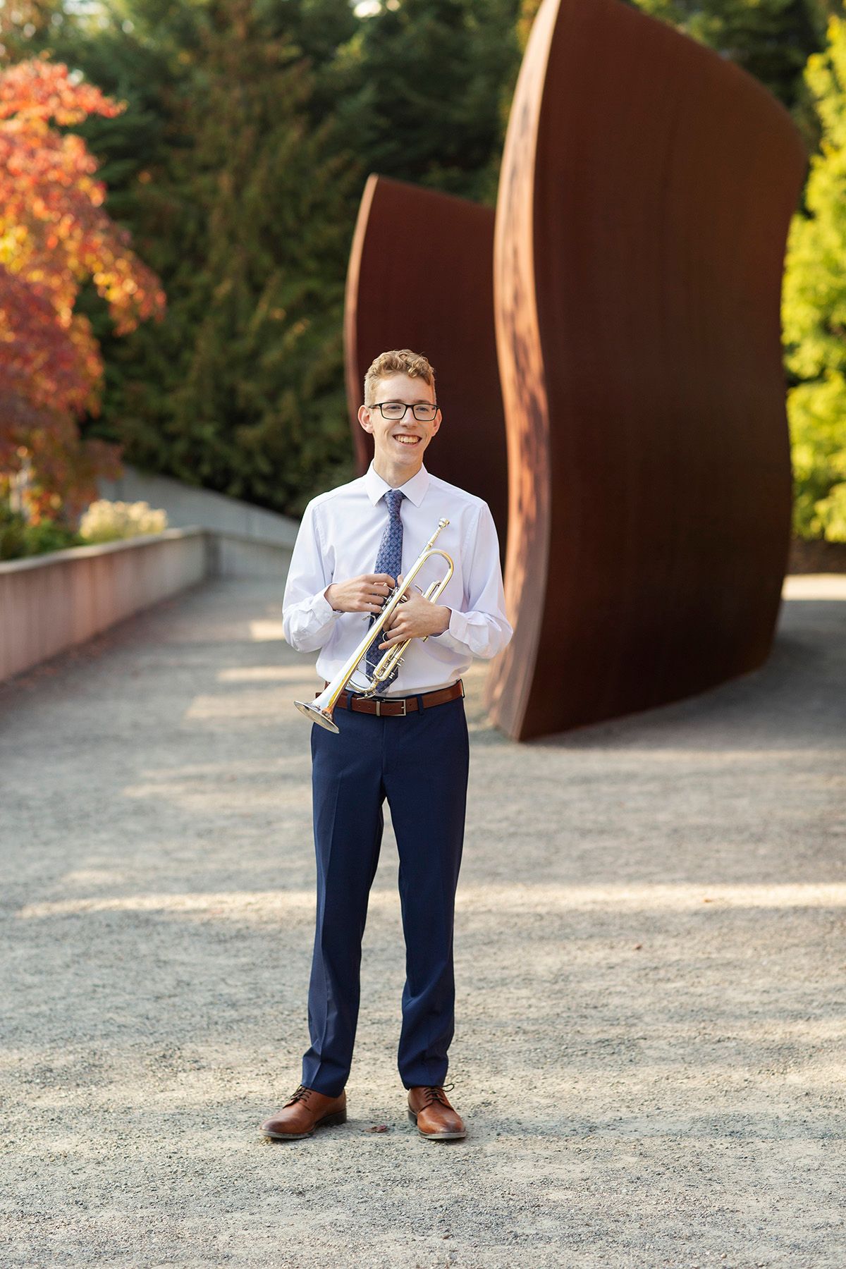 Young musician in dress shirt and blue pants holds a trumpet outdoors near a rustic metal sculpture.