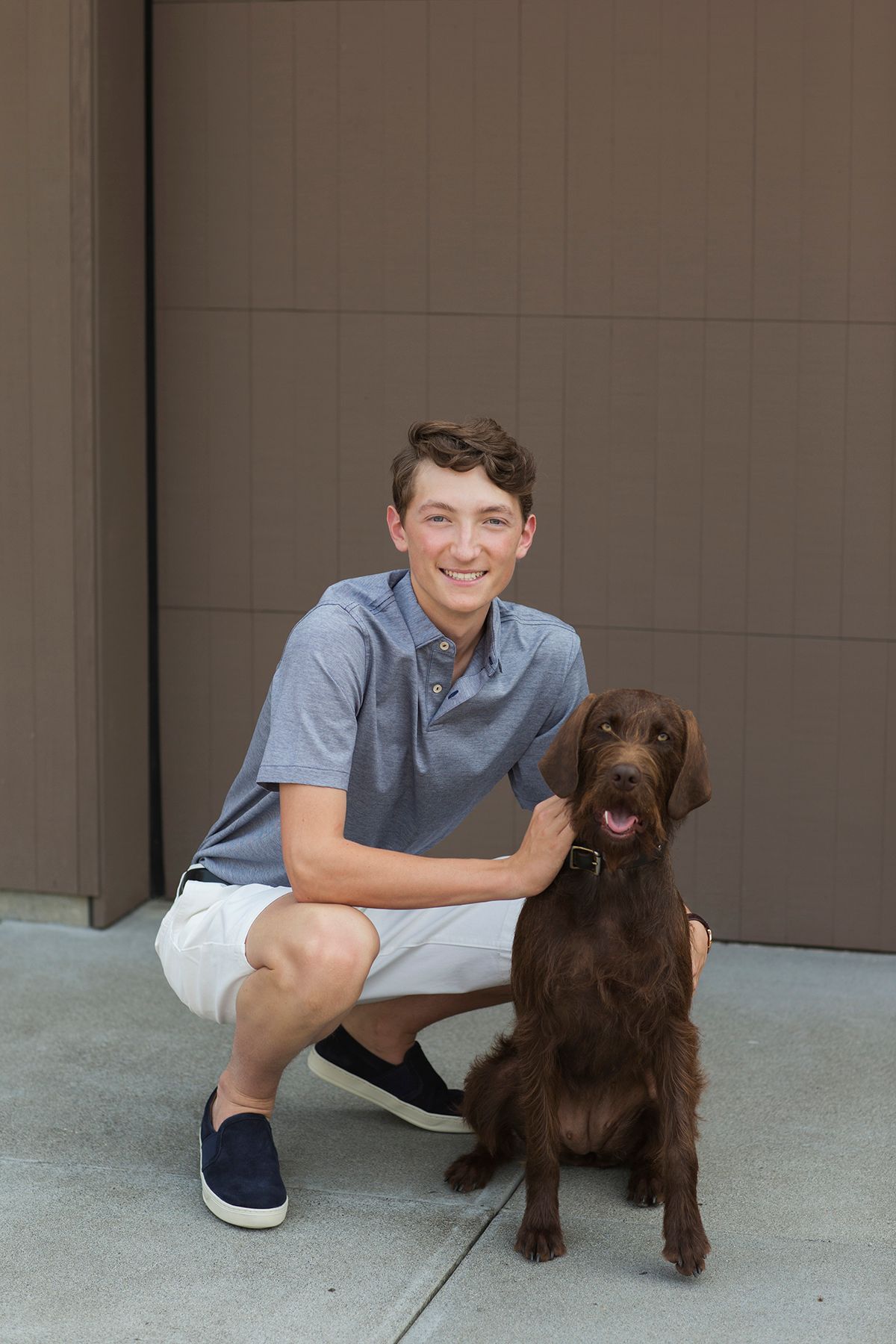 Smiling teenage boy crouching beside a chocolate Labrador dog in front of a modern gray building.