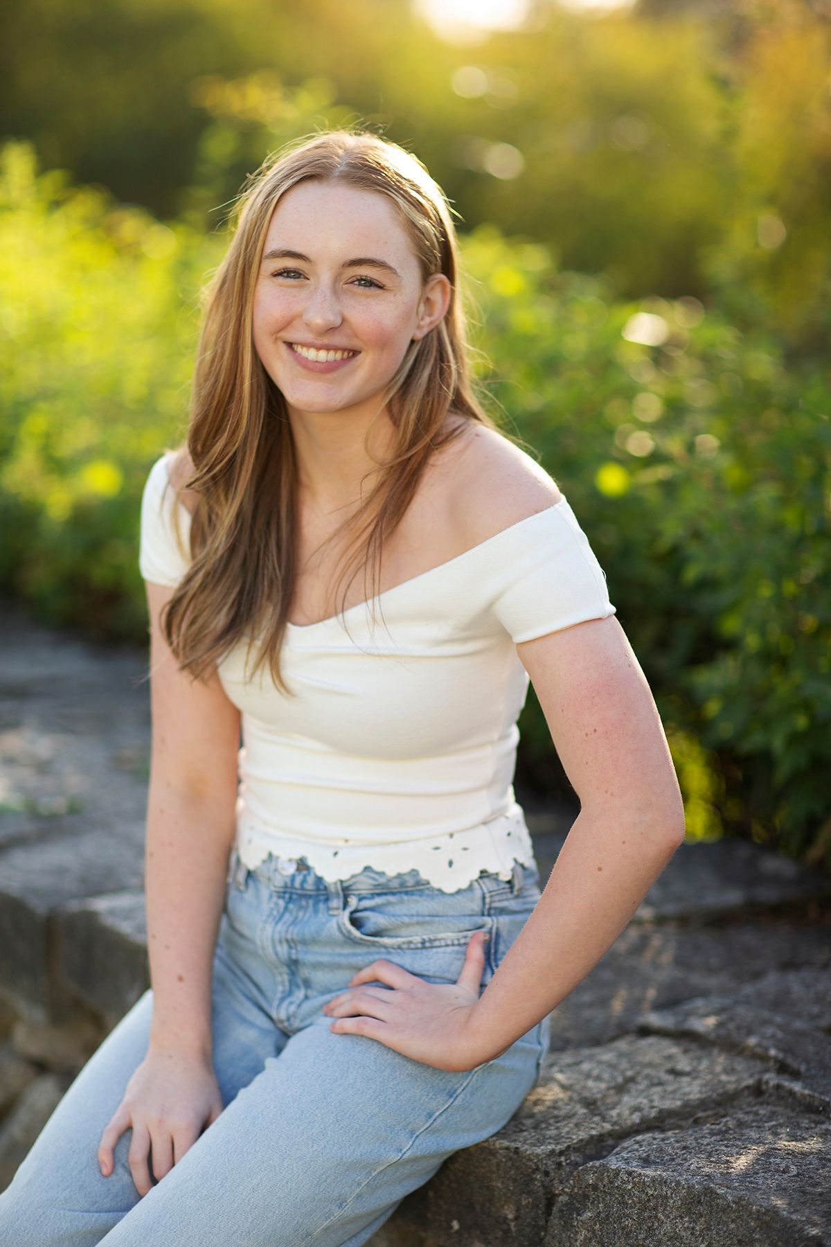 Smiling young woman in white off-shoulder top and jeans sitting on stone wall outdoors in golden sunlight.