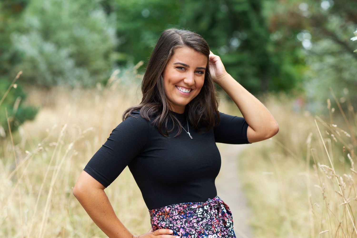 Cheerful brunette woman in black top and floral skirt posing outdoors in a golden grass field.