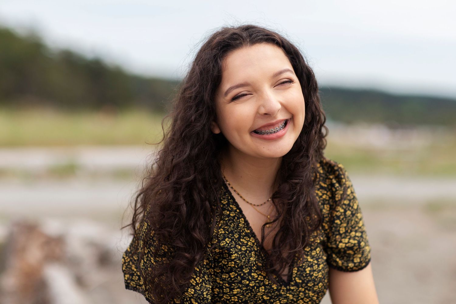 Smiling teenage girl with braces and curly hair in floral dress at beach.