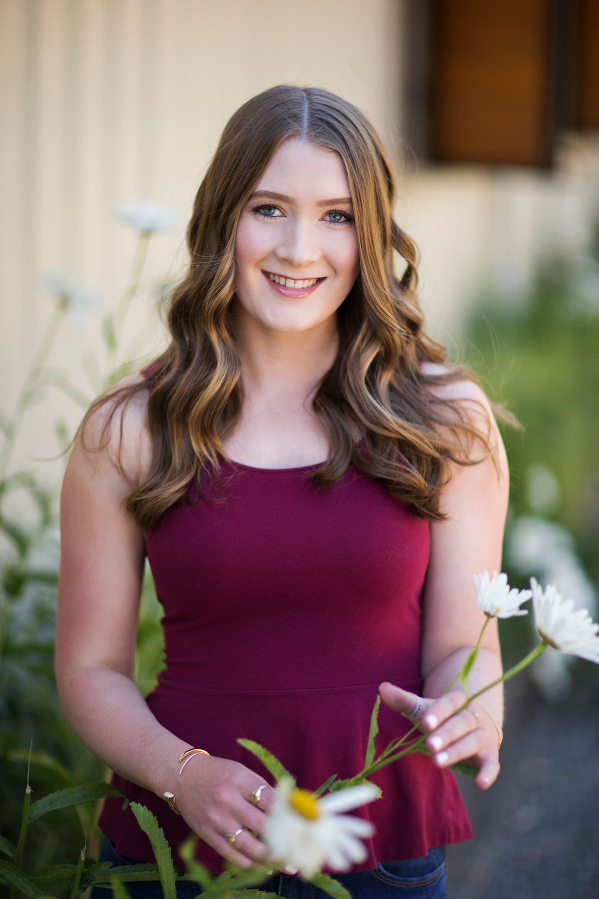 Young woman in burgundy top smiling outdoors holding white daisies with wavy brown hair in natural light.