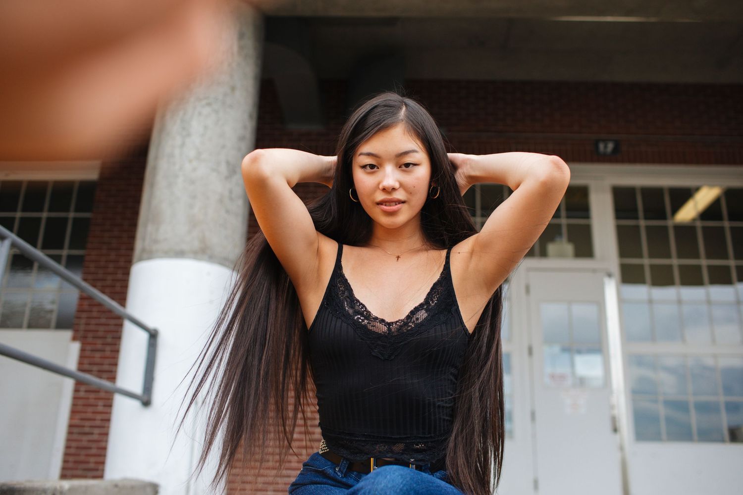 Young woman with long dark hair posing with hands behind head outdoors near brick building.
