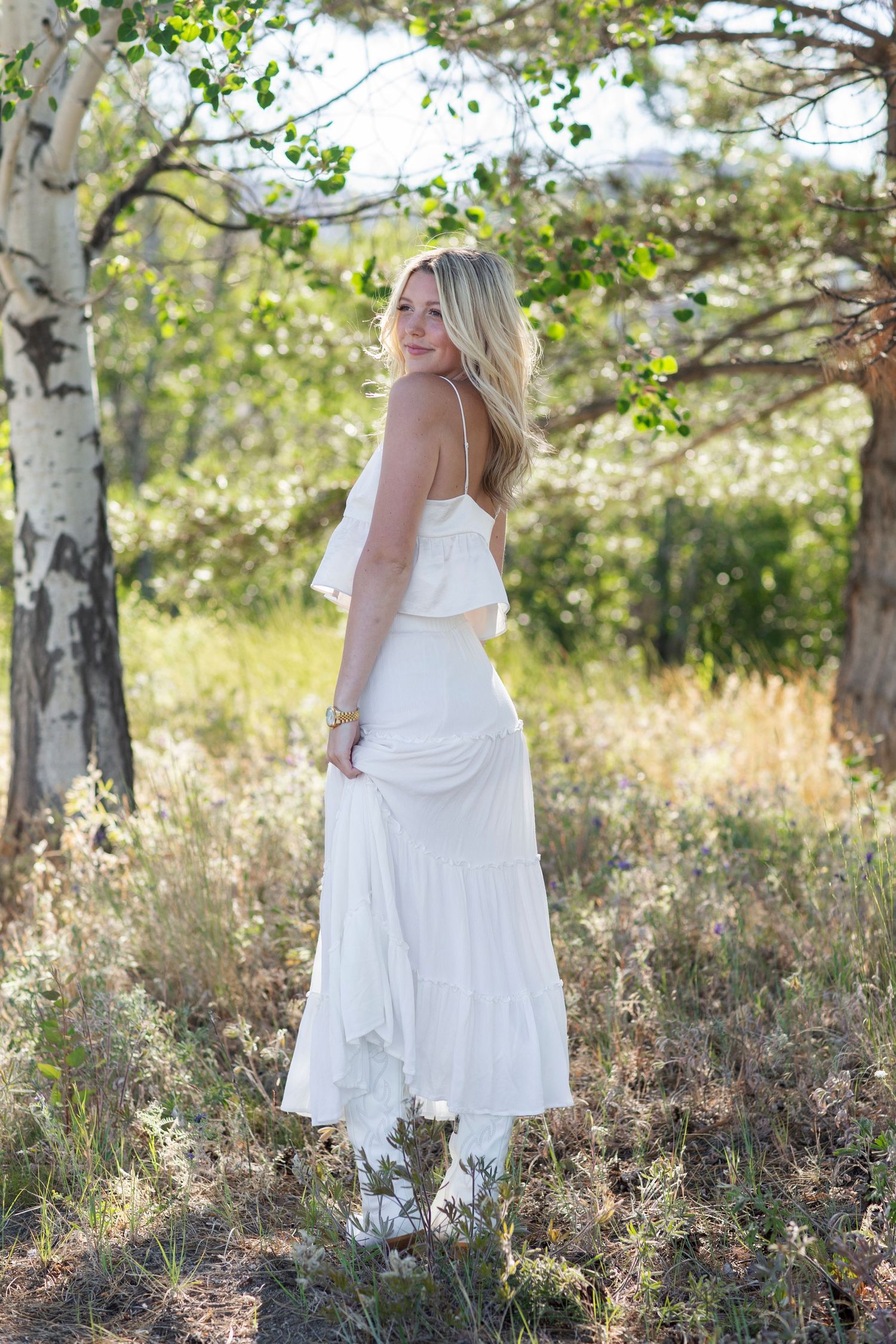 Blonde woman in white strapless ruffle dress looking back over shoulder while standing among birch trees.