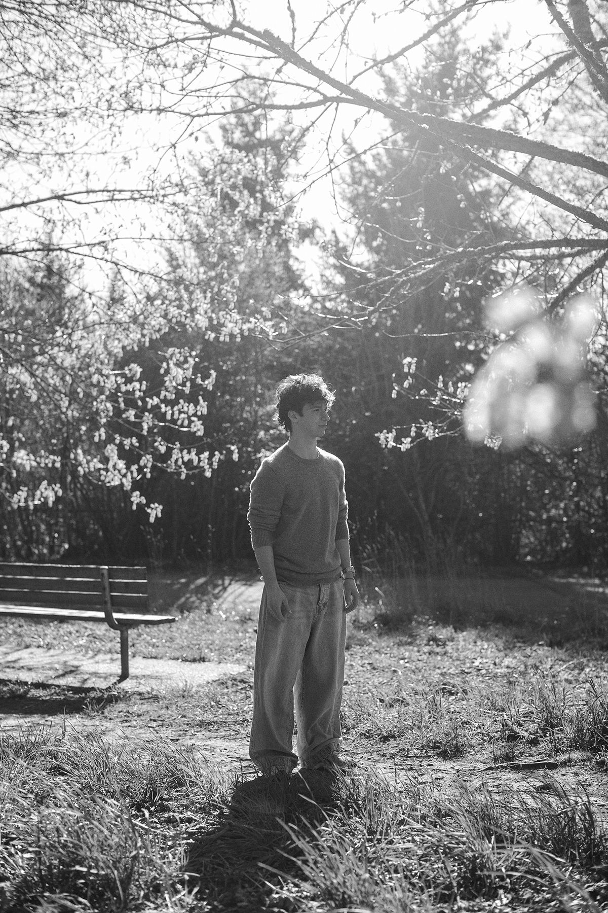 Black and white photo of a young man standing in a sunlit park surrounded by trees and a bench in early spring.